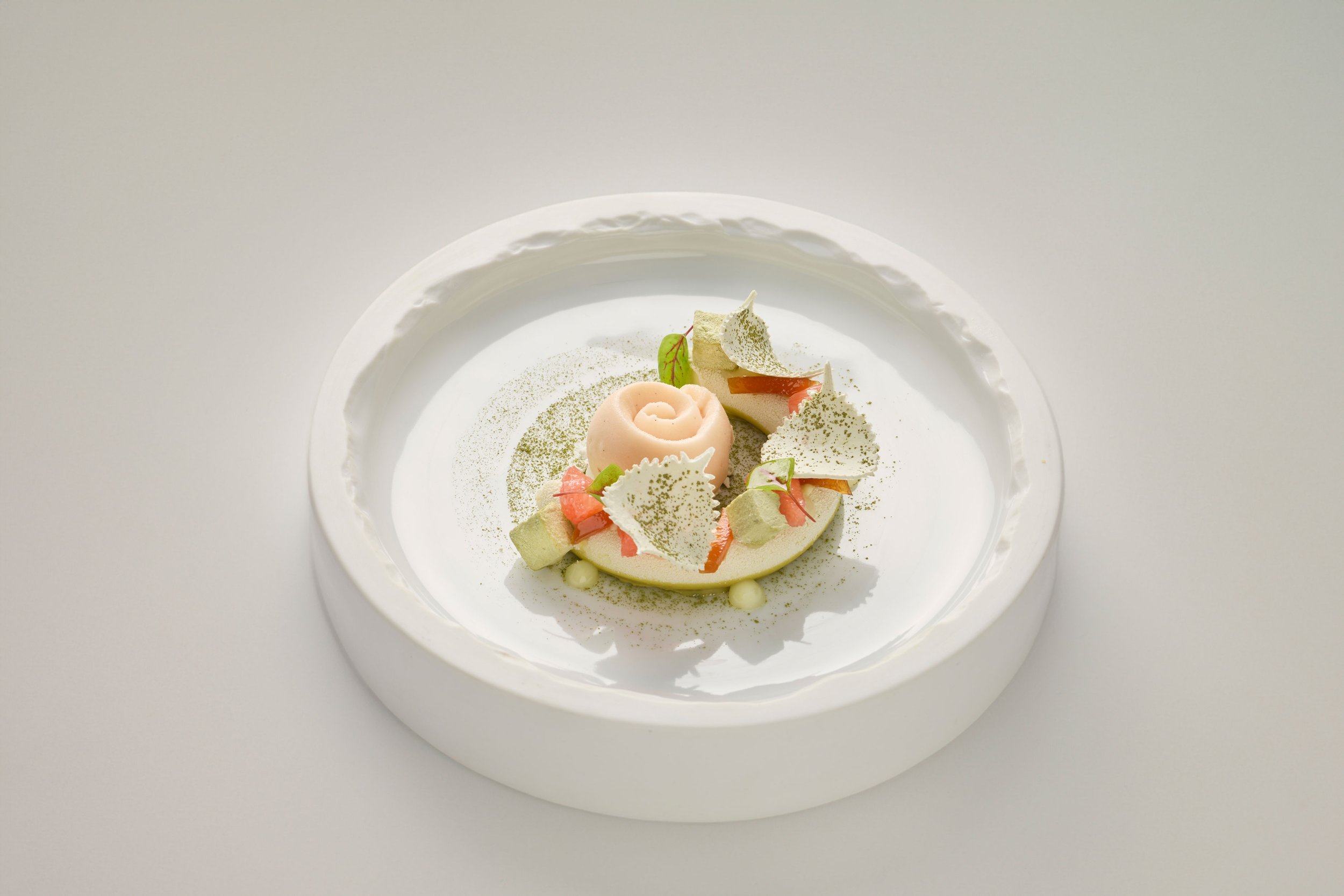 Dessert sophistiqué avec un motif de rose, décoré de feuilles, de mousse verte, et de croustillants blancs, présenté sur une assiette blanche élégante.