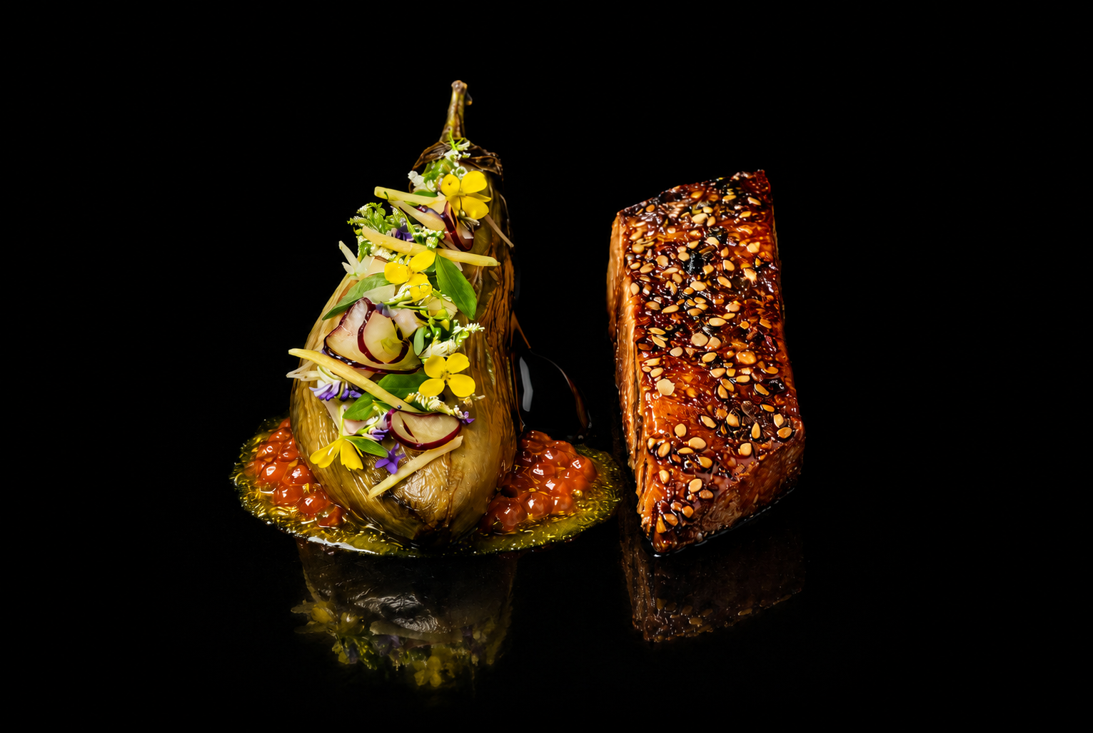 Deux plats gastronomiques sophistiqués, un légume farci décoré de fleurs et de fines herbes, et un bloc de viande glacée recouvert de graines de sésame, présentés sur un fond noir.