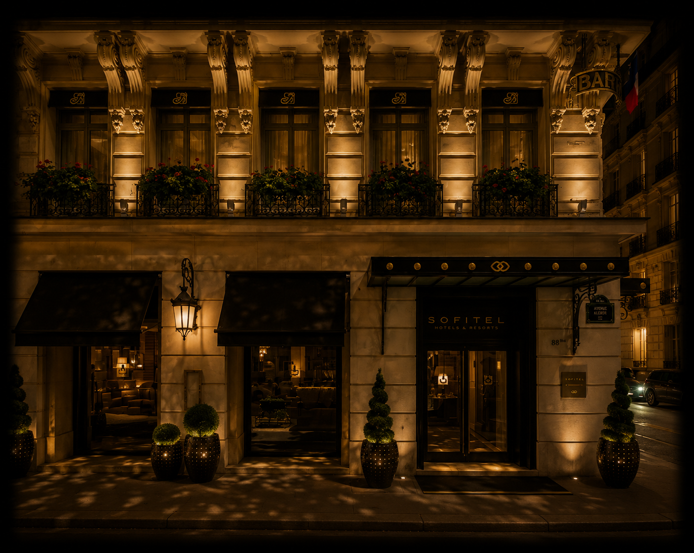 Façade lumineuse d'un hôtel Sofitel la nuit avec des fenêtres éclairées, des plantes en pot devant l'entrée, un lampadaire et des balcons avec des fleurs.