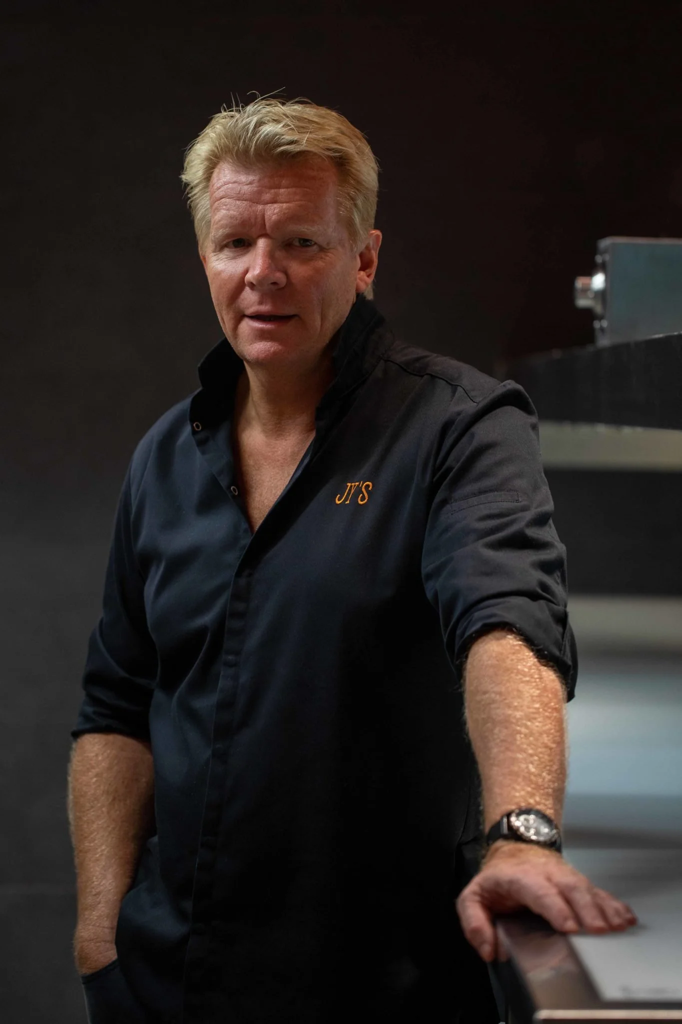 Un homme avec des cheveux blonds et une veste noire avec l'inscription JJ'S, debout près d'un comptoir en noir, dans une pièce sombre.