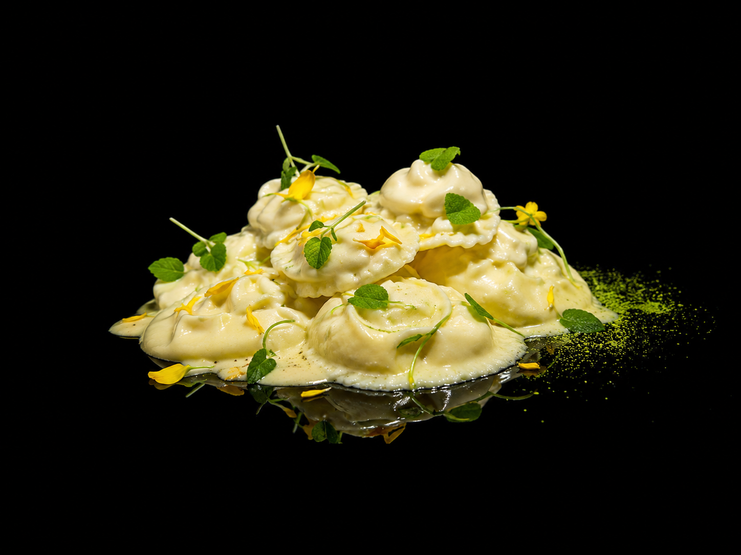 Assiette de raviolis garnis de sauce blanche, décorée de feuilles de menthe et de petites fleurs jaunes, sur un fond noir.