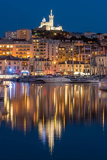 Vue nocturne d'un port avec des bateaux amarrés et des bâtiments résidentiels, avec une colline en arrière-plan où se trouve une statue de la Vierge Marie surplombant la ville.