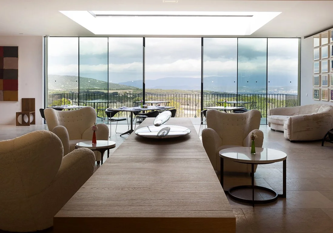 Salon avec grands fenêtres offrant une vue sur la nature et un ciel nuageux, mobilier moderne avec des fauteuils beiges et des tables basses, décor épuré, quelques fleurs dans des vases à côté des fauteuils.