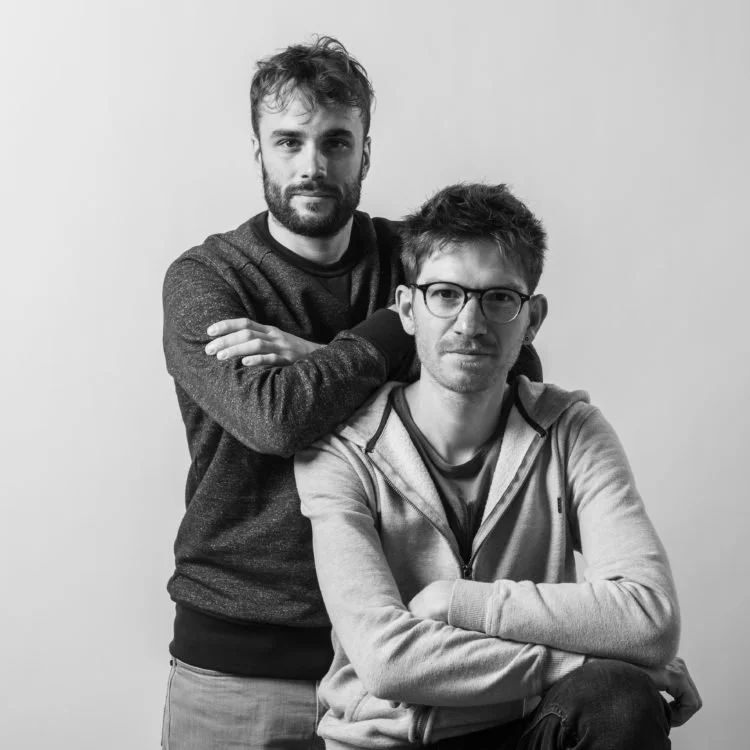 Deux hommes posent ensemble, un debout derrière et l'autre assis devant, fond blanc, en noir et blanc.