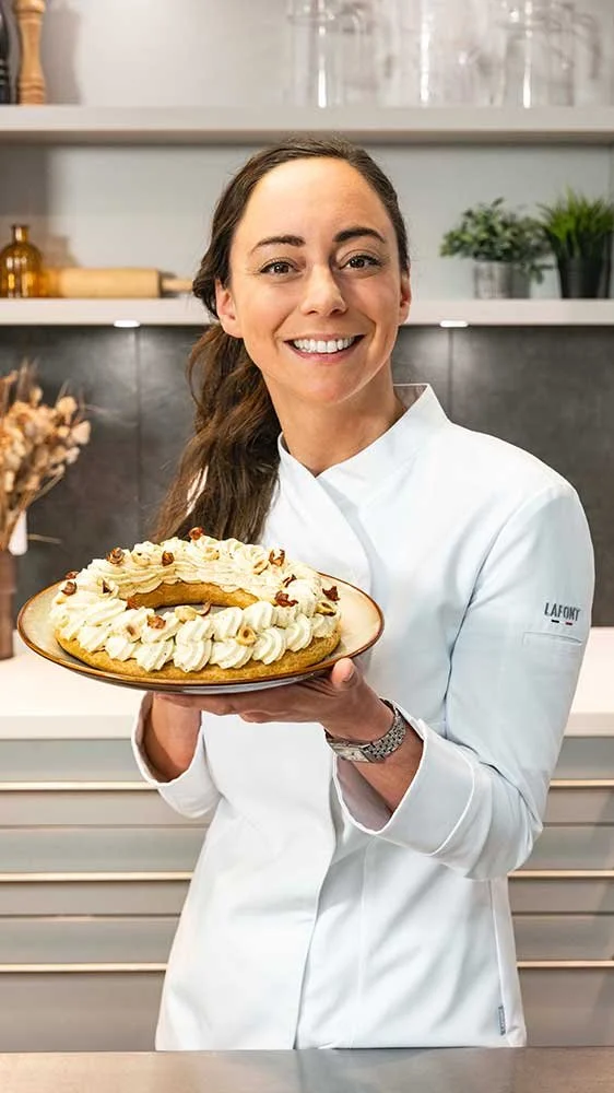 Femme chef en uniforme blanc tenant un gâteau décoré avec de la crème et des noix dans une cuisine moderne.