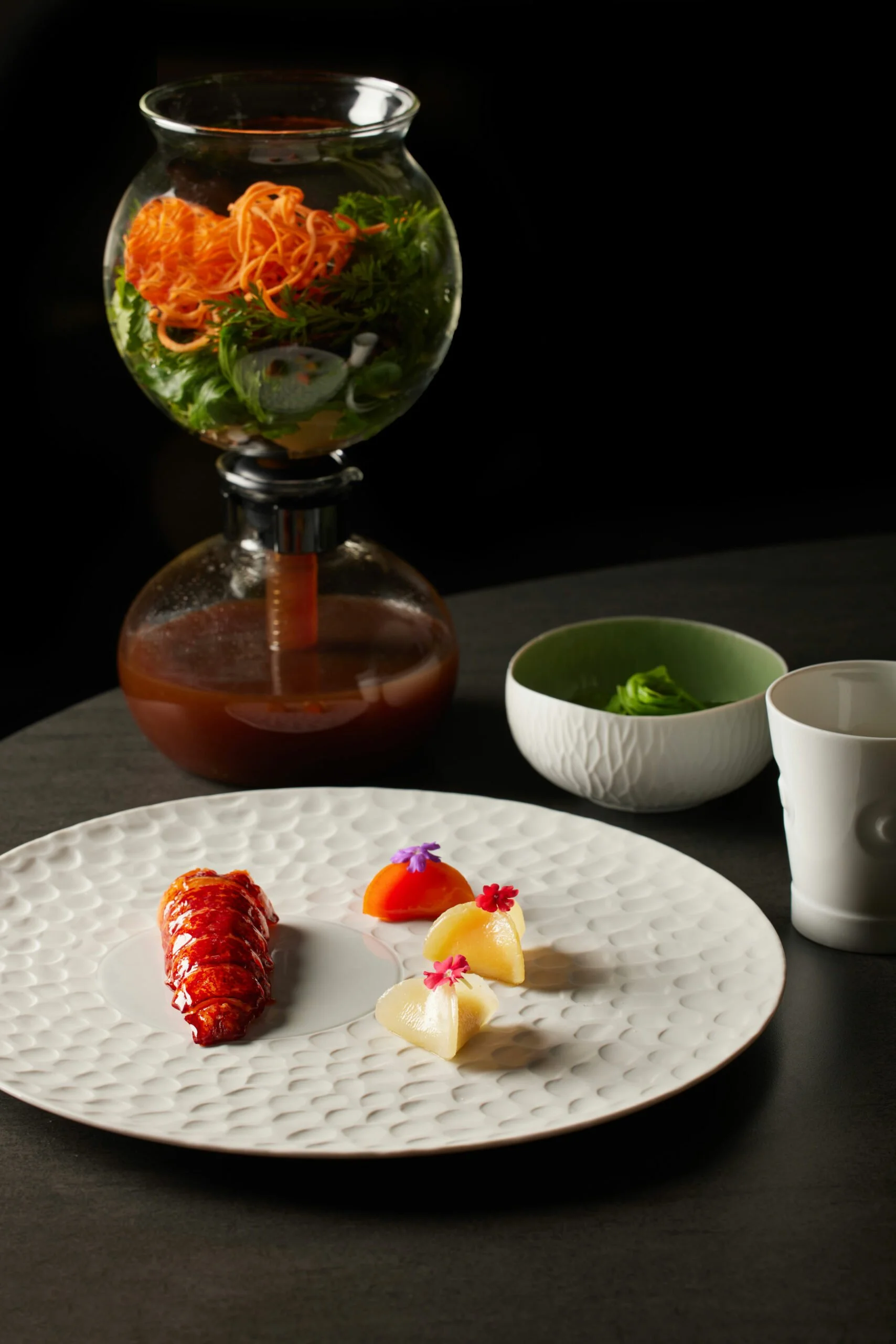Assiette de sushi avec homard, accompagnée de petites salades et de garnitures décoratives, avec un service à thé en porcelaine blanche à côté, devant une fontaine à lave de salade.