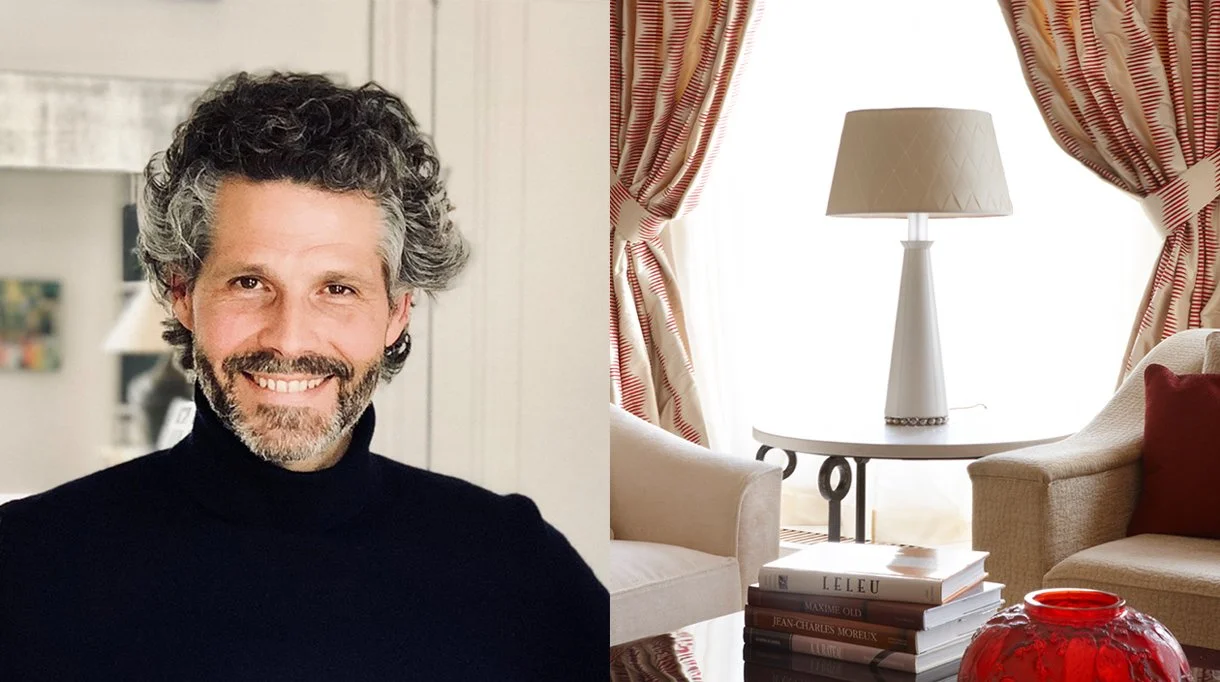 Une photo d'un homme souriant avec des cheveux gris et une barbe, portant un pull noir, dans un intérieur lumineux avec des meubles beige et des rideaux à rayures rouges et blanches.