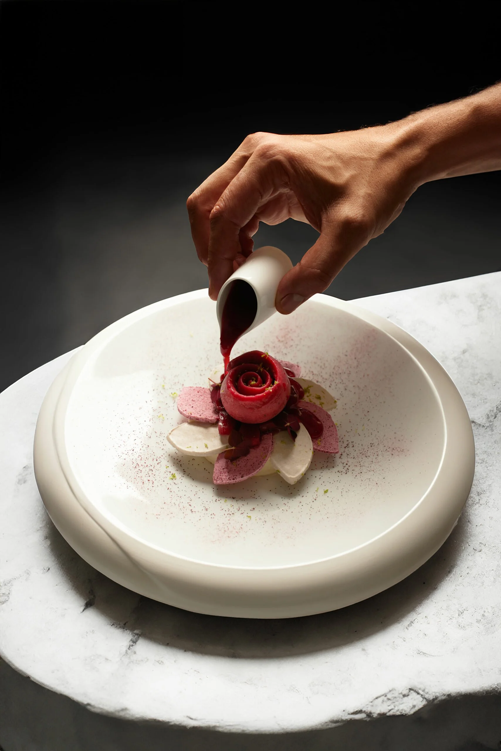 Une main verse du coulis rouge sur une présentation de dessert en forme de rose, avec des pétales colorés et une poudre fine sur une assiette blanche.