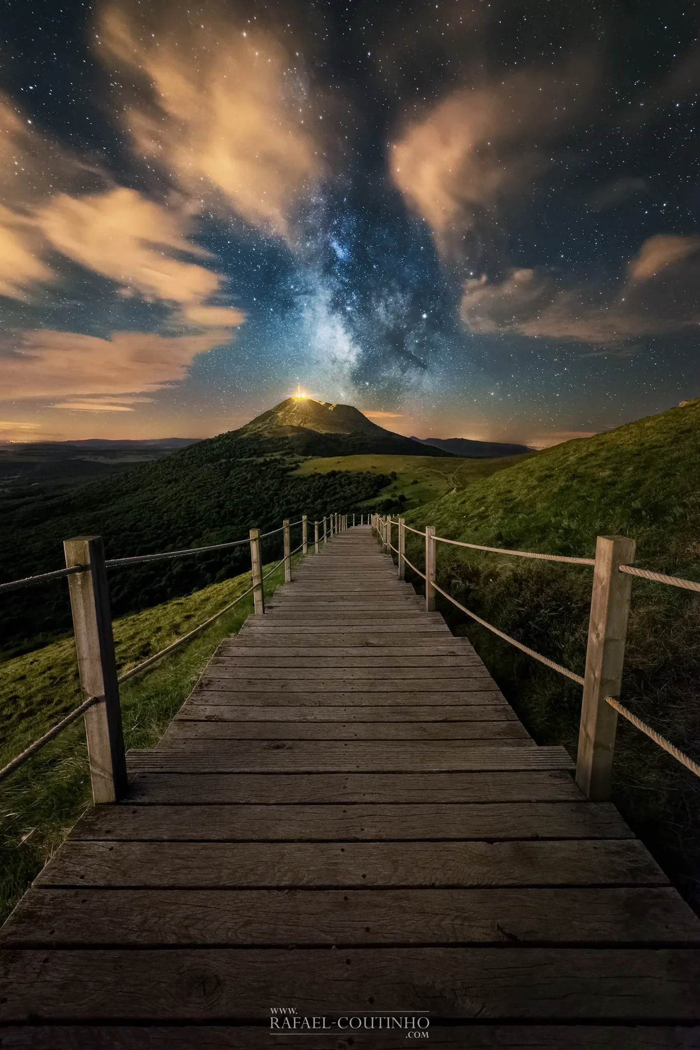 Un chemin en bois menant vers une colline avec un ciel étoilé et la voie lactée, des nuages orangeâtres dans le ciel, et une lumière brillante au sommet de la colline.
