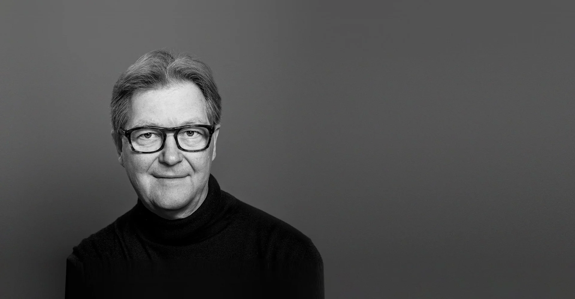 Portrait en noir et blanc d'un homme portant des lunettes et un pull noir, face à la caméra, avec un sourire subtil.