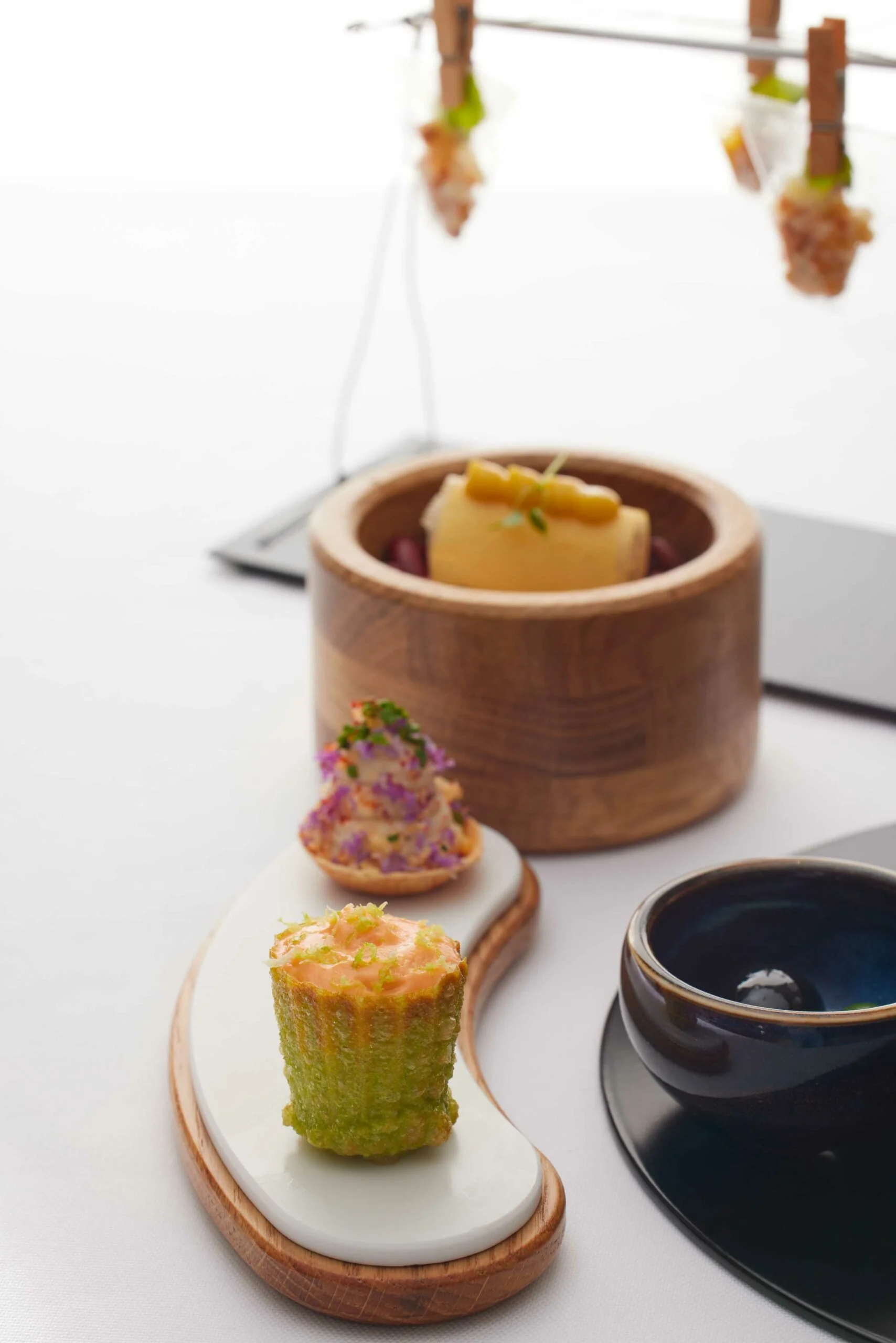Assortiment d'amuse-bouches présentés sur une planche en bois, incluant une coupe de caviar, un muffin aux œufs de saumon et un rouleau d'algues, avec un bol noir à côté, le tout sur une nappe blanche.