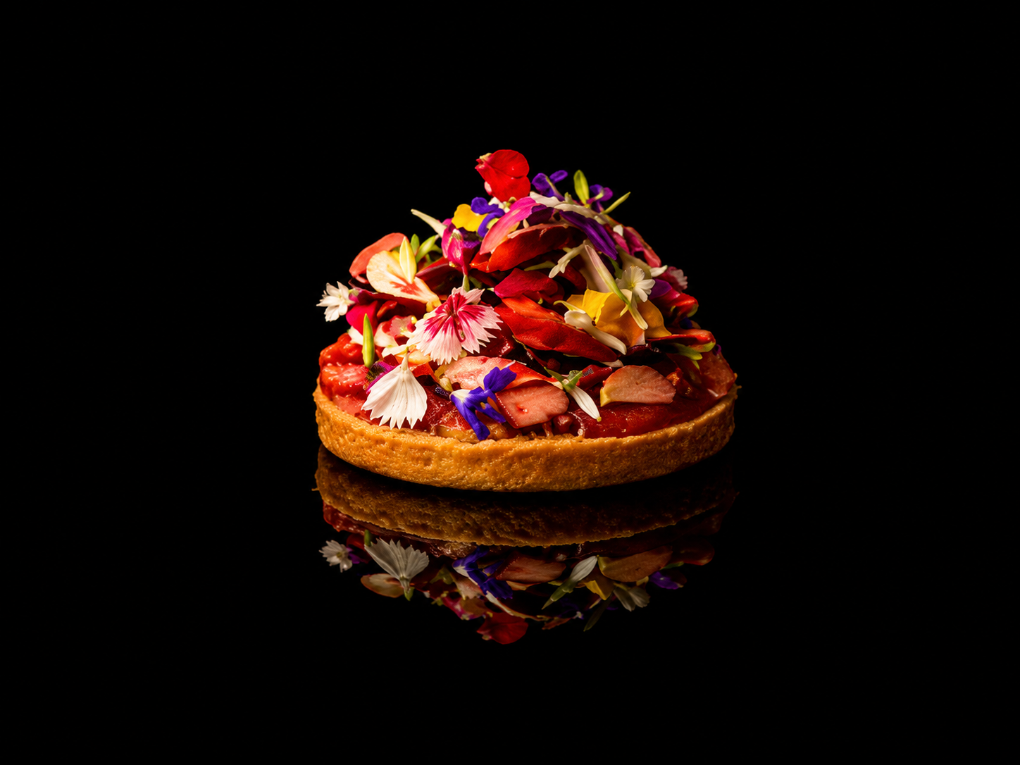 Une tarte aux fruits décorée avec des fleurs comestibles sur un fond noir, avec une réflexion miroir