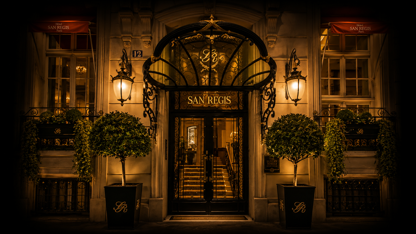 Entrée élégante d'un hôtel nommé San Régis à Paris, décorée avec une porte en fer forgé, deux lampes murales, deux grands pots avec des arbustes, et un plafond lumineux visible à travers la porte, ambiance chaleureuse et luxueuse.