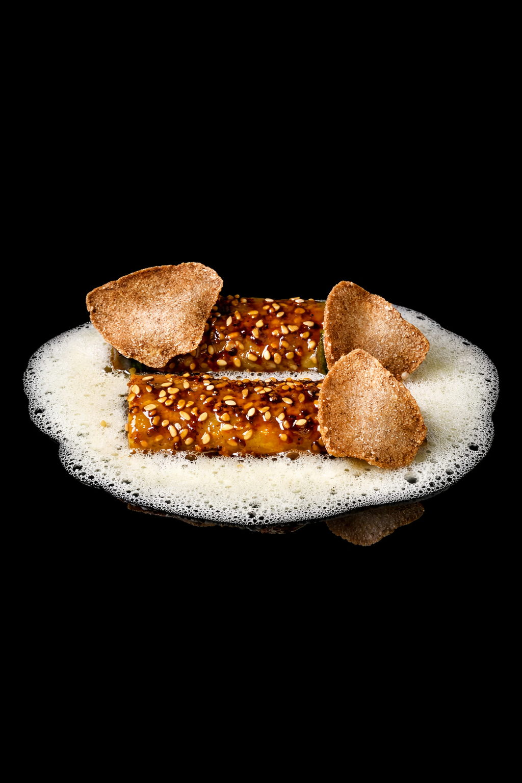 Deux rouleaux de sushi recouverts de sésame, entourés de chips frites, avec une mousse blanche autour, sur un fond noir