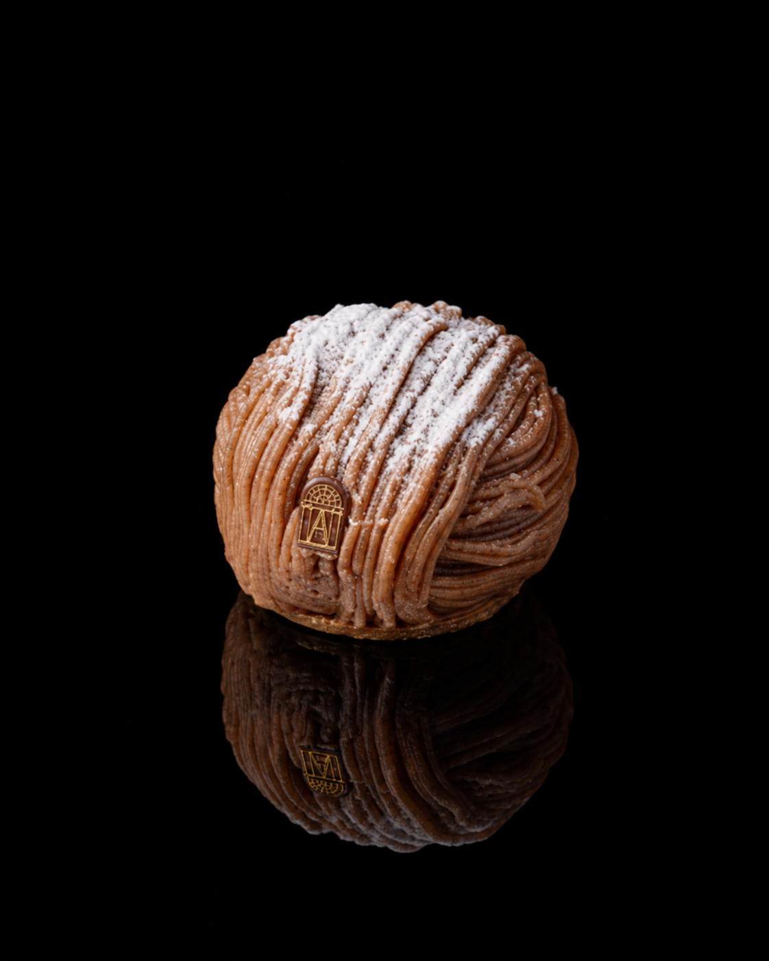 Dessert en forme de boule, avec une surface en filaments de couleur beige, saupoudré de sucre glace, avec un petit badge doré sur le devant, posé sur une surface noire avec reflection.