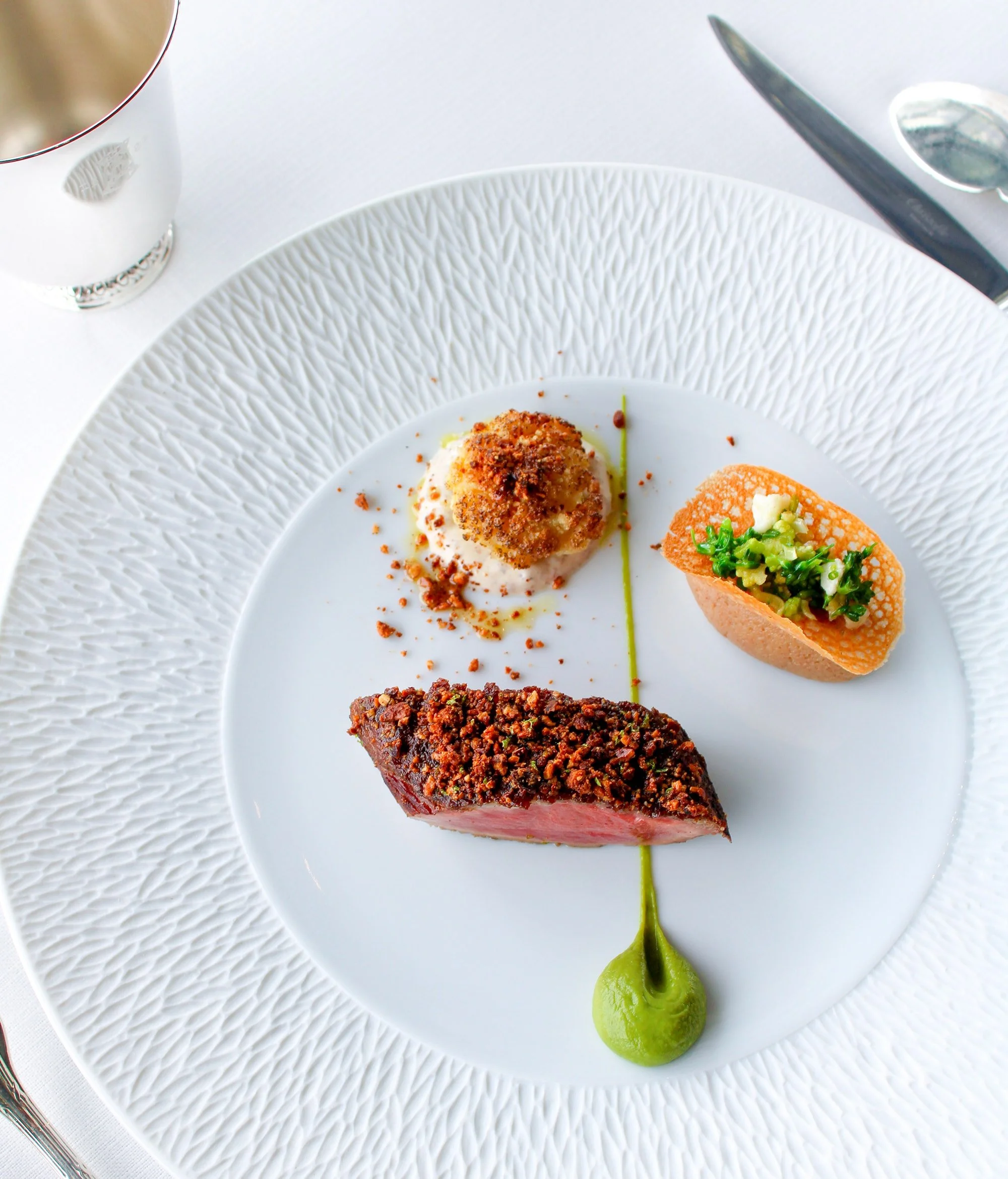 Plate de viande rouge nappée d'une croûte croustillante, accompagnée de terre cuite de purée et d'une quenelle de vert, avec un petit jaune en haut.