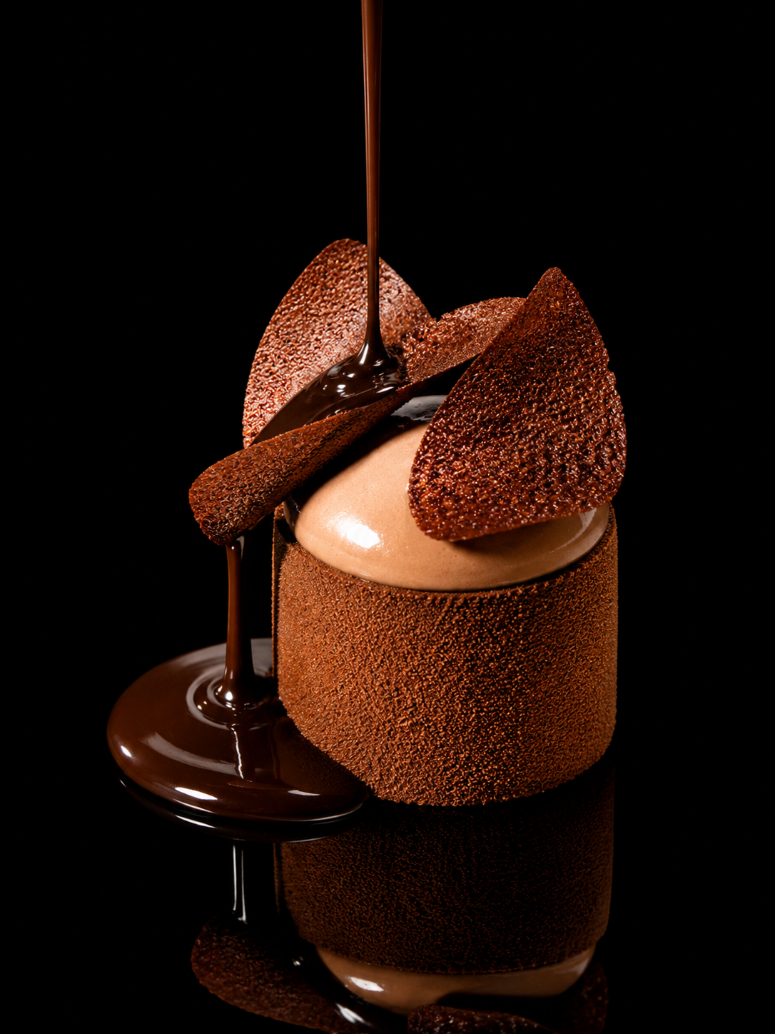 Un dessert au chocolat avec une coque en chocolat, des morceaux de chocolat croquants, et du chocolat liquide coulant dessus, contre un fond noir.