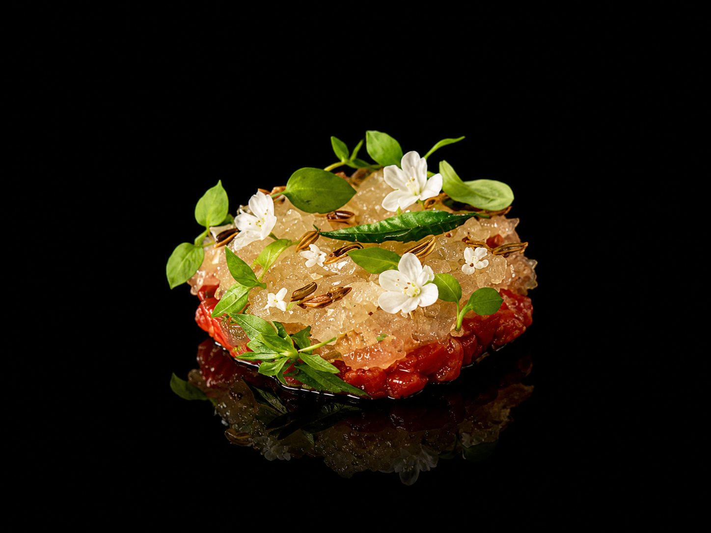 Présentation culinaire d'un tartare de poisson ou de viande grillé, décoré de fleurs blanches, de feuilles vertes et de graines sur un fond noir