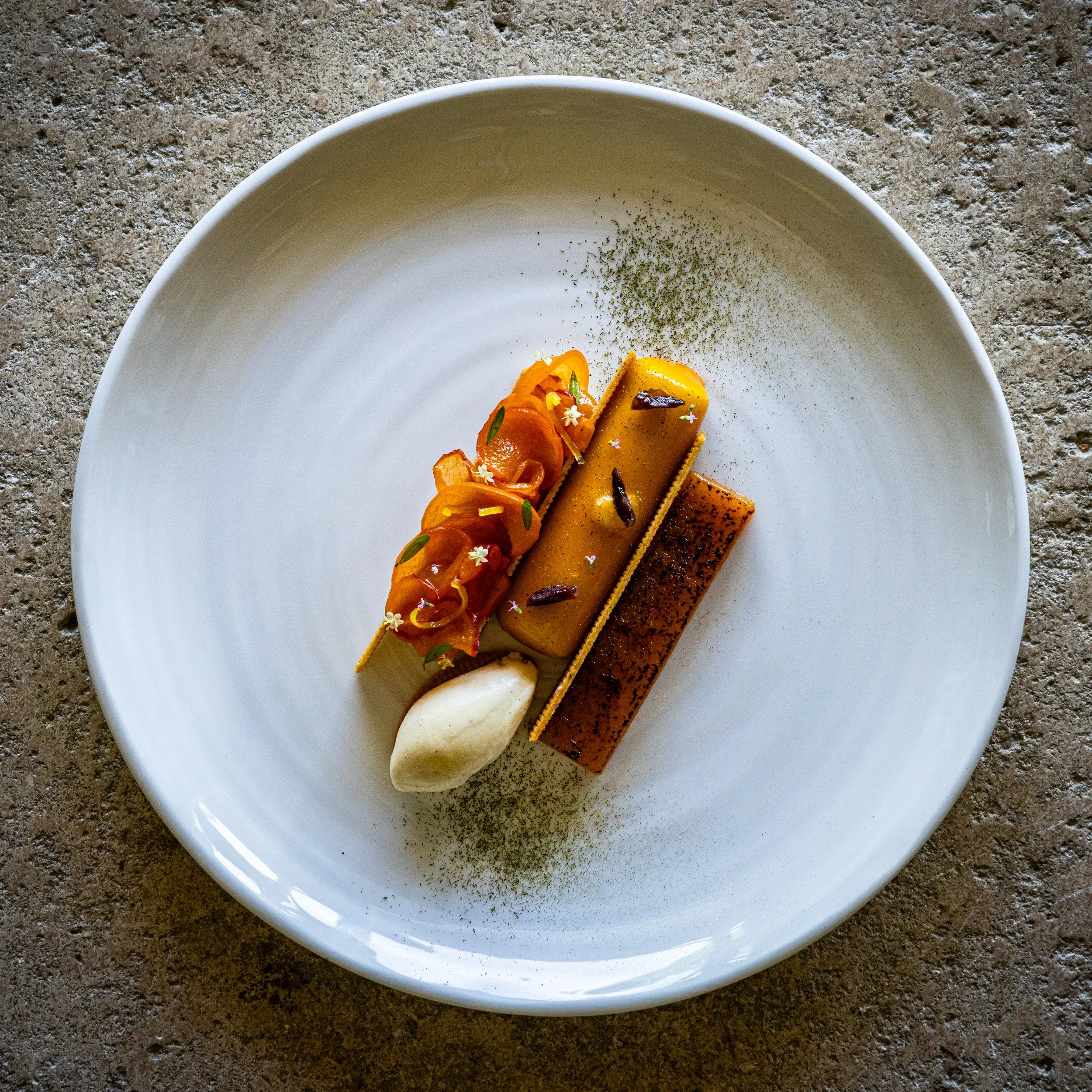 Dessert élégant avec deux rectangles de gâteau, une petite quenelle de crème, et des décorations florales et en herbes sur une assiette blanche.