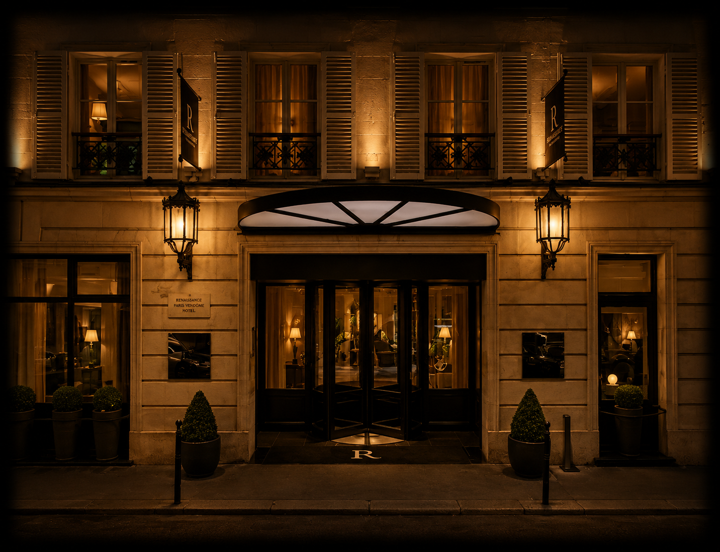 Façade d'un hôtel élégant avec une enseigne « Renaissance Paris Vendôme Hotel » éclairée, fenêtres avec des rideaux et lampes intérieures, deux lampadaires en façade, plantes dans des pots et porte d'entrée centrale sous un auvent lumineux, ambiance chaleureuse nocturne.
