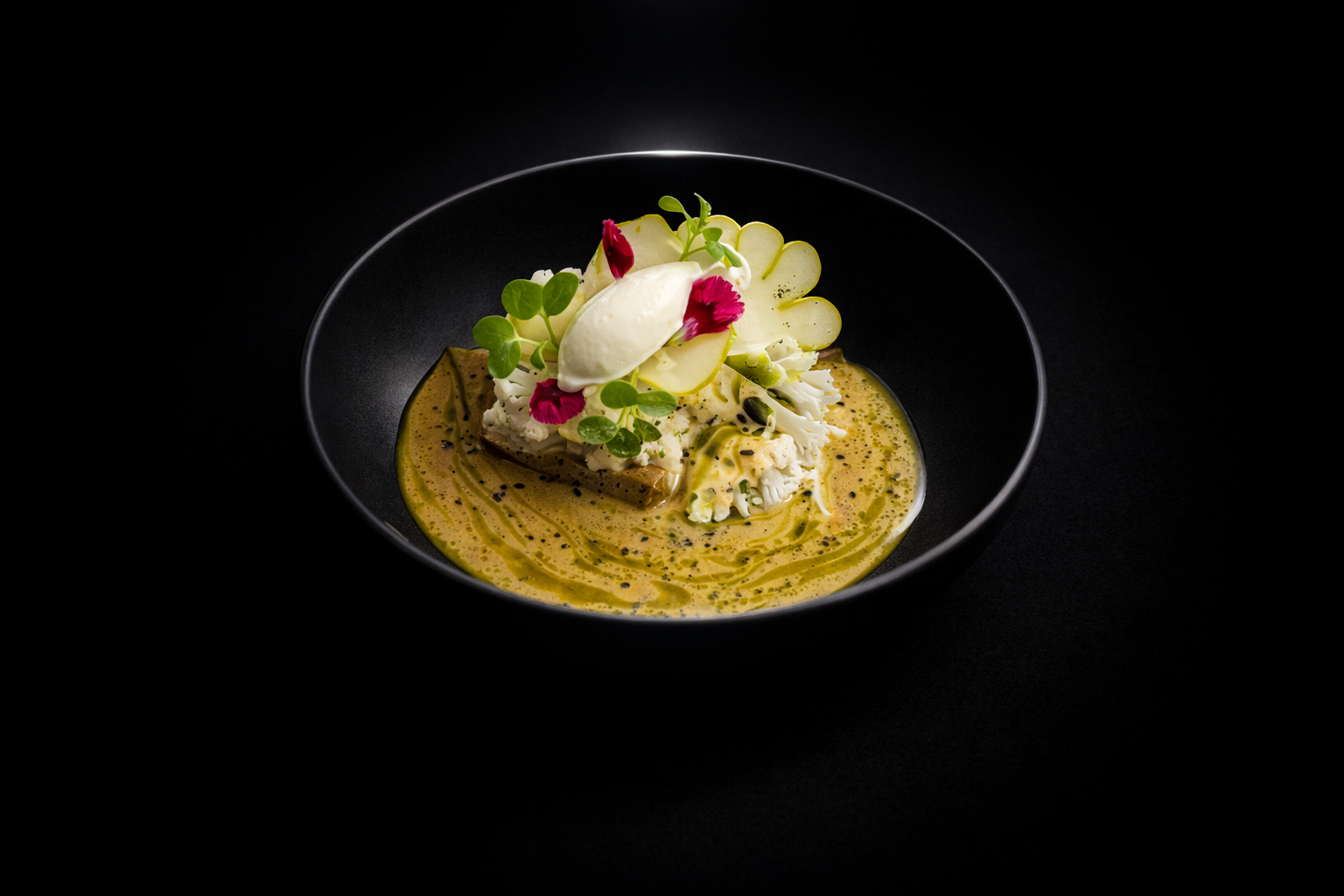 Assiette noire avec un plat gastronomique comprenant une sauce verte, des légumes en décoration, une quenelle de crème blanche et des fleurs comestibles roses et vertes.