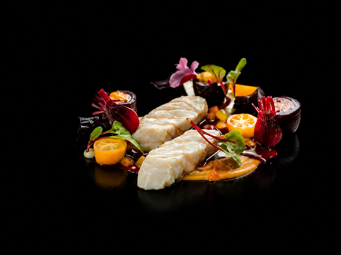 Filet de poisson blanc avec une sauce entourée de légumes comme citrons, betteraves, carottes, plusieurs feuilles vertes et décoration florale sur un fond noir.