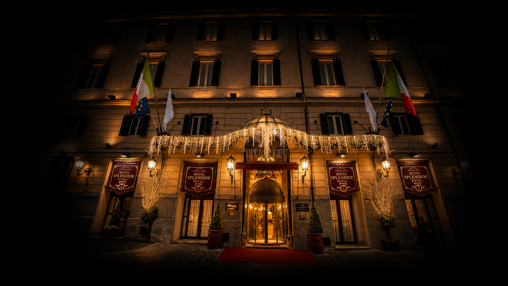 Façade d'un hôtel éclairée avec des décorations de Noël, drapeaux italien et européen, et une entrée avec un tapis rouge.
