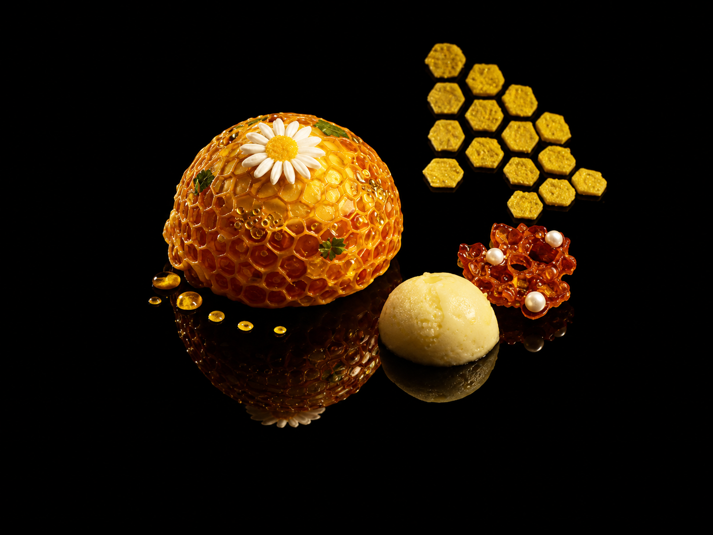 Une collection de miels et de bonbons décoratifs présentés sur un fond noir, comprenant un dôme de miel avec une marguerite en décoration, des cristaux de miel, des bonbons en forme de hexagones, une boule de glace ou de fromage, et un éclat de miel avec des perles.