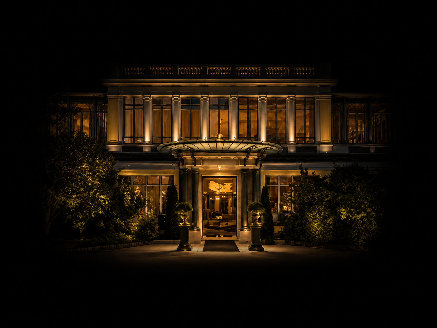 Façade d'un bâtiment élégant illuminé la nuit avec des lumières chaudes, grande entrée, colonnes et jardins bien entretenus.