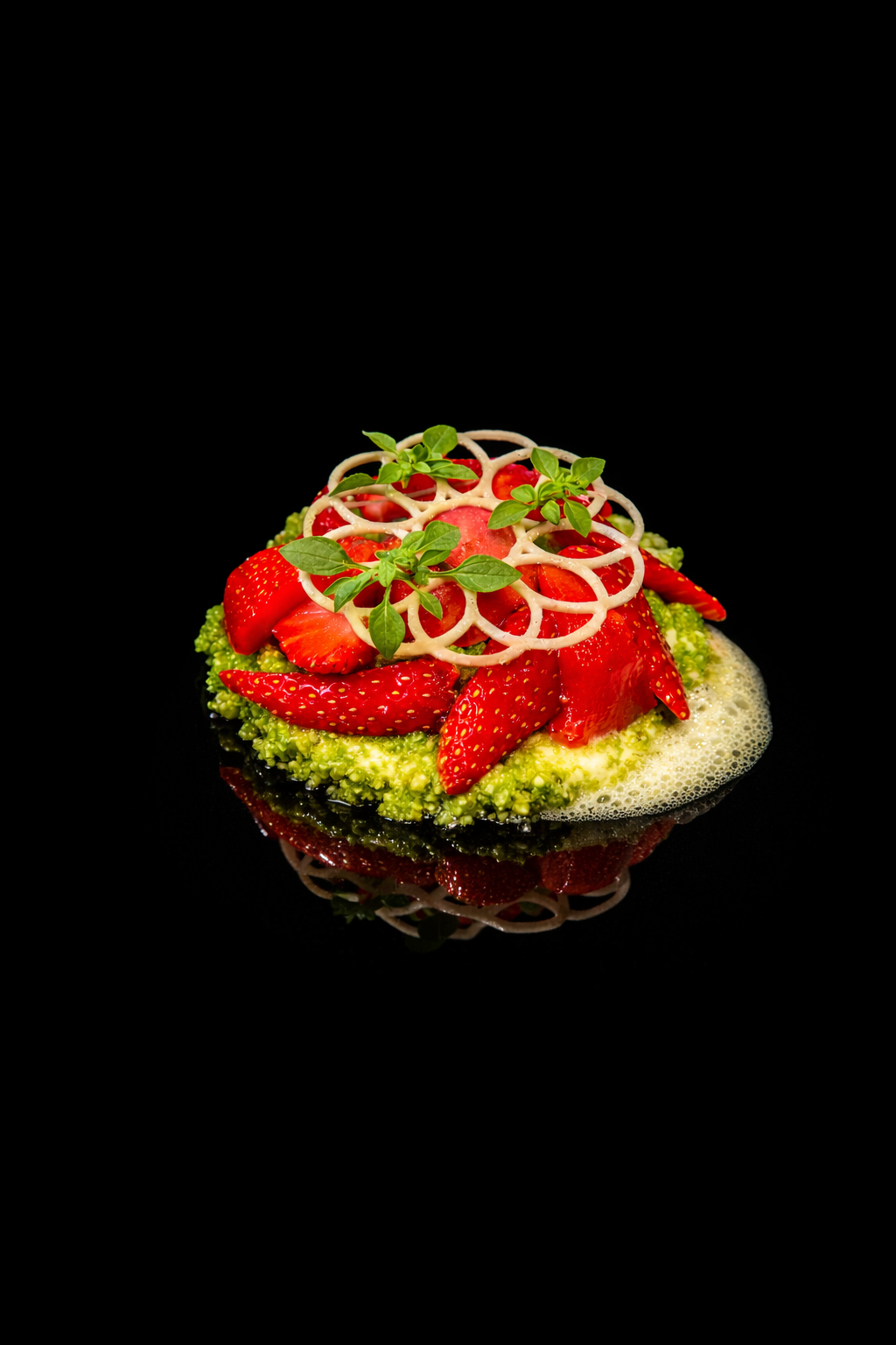 Dessert avec des fraises coupées, de la réduction de basilic, une décoration en croustillant et un coulis vert, présenté sur fond noir.