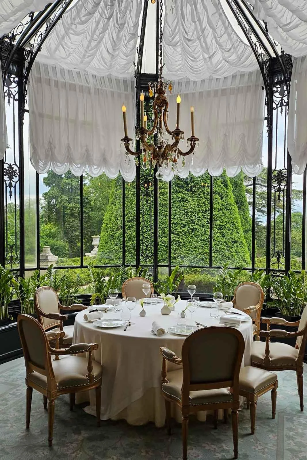 Salle à manger élégante avec une table ronde drapée de blanc, entourée de chaises en bois et en tissu beige, ornée de verres à vin, assiettes et napperons. Végétation luxuriante vue à travers de grandes fenêtres en verre, sous un plafond décoratif blanc avec chandelier en or.