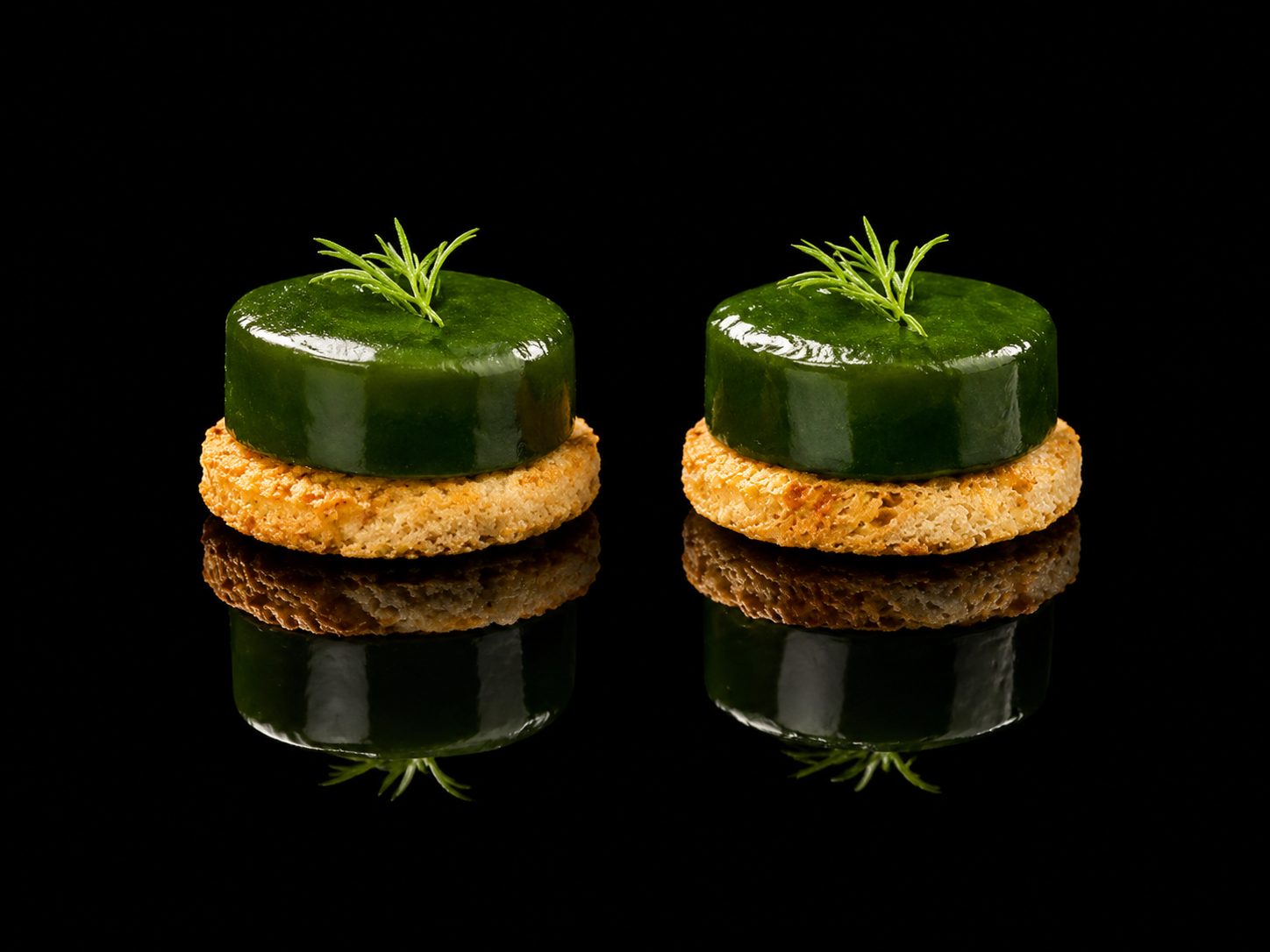 Deux petits amuse-bouches avec un croûton, une rondelle de courgette glacée, et une petite branche de romarin, avec une reflection sur une surface noire.