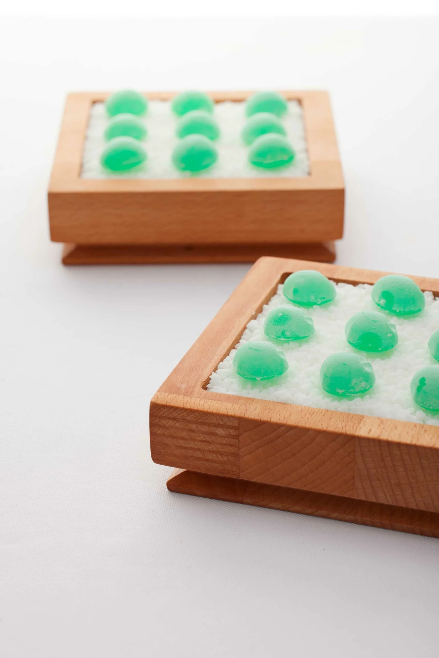 Deux boîtes en bois contenant des savons ou cristaux verts dans un coussin de sel ou de sable blanc.