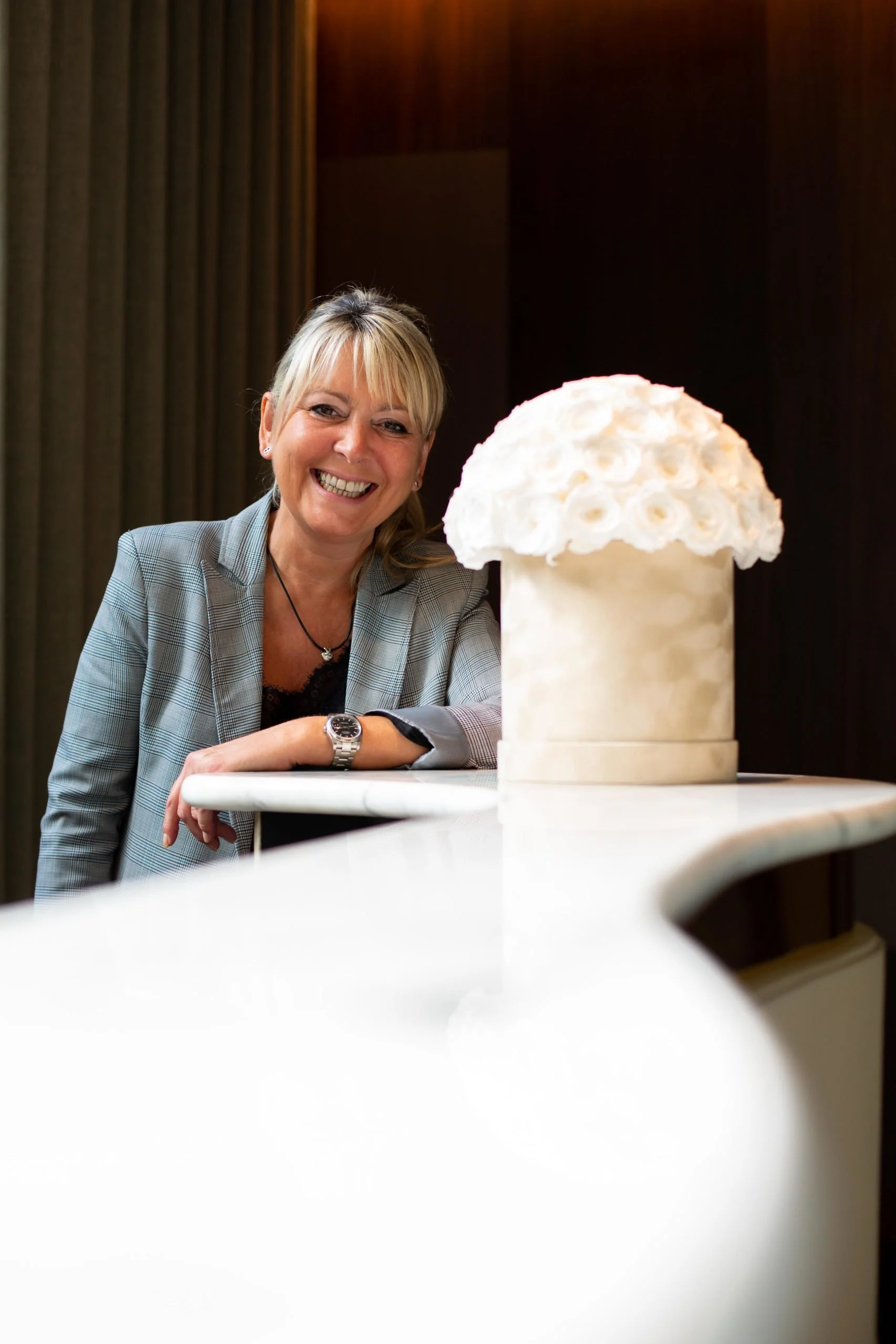 Une femme souriante avec une veste grise pose près d'une sculpture blanche en forme de champignon ou de nuage sur une table beige.