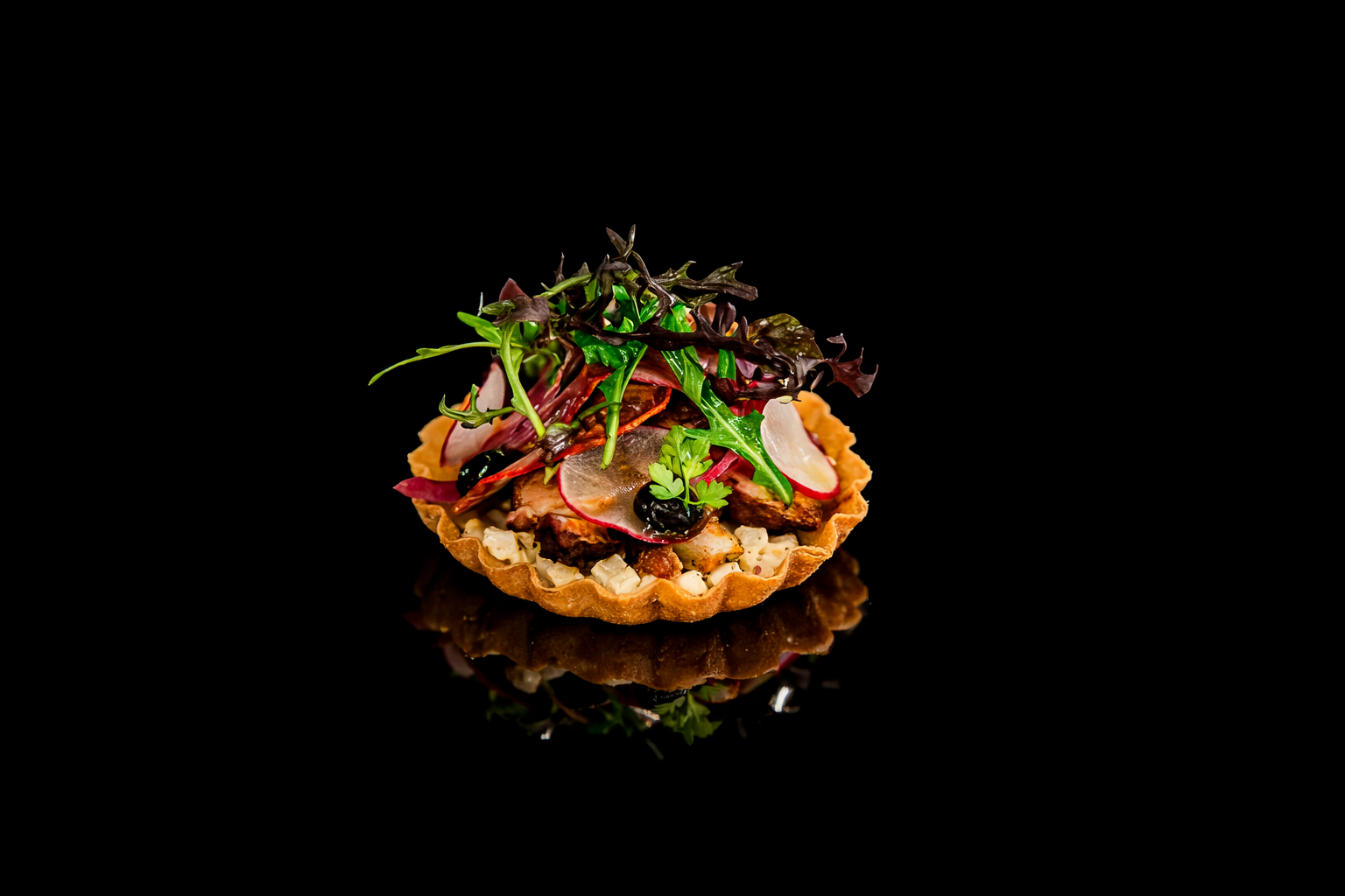 Tartelette salée garnie de salade, radis, olives et herbes, avec un fond noir.