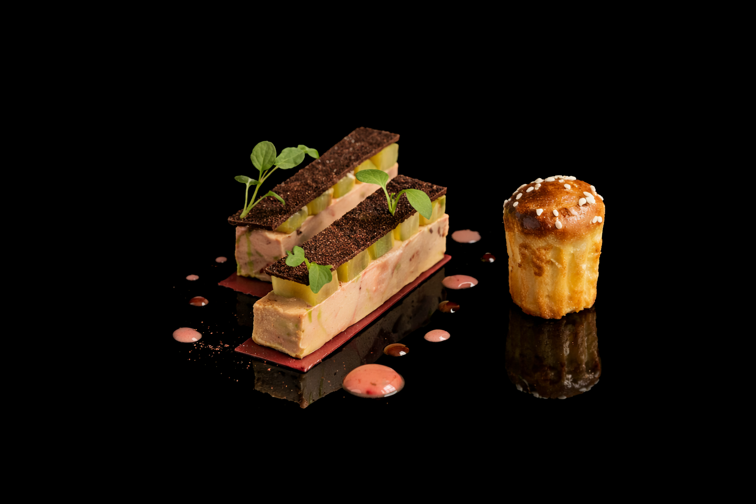 Gourmet dessert plate with layered terrine garnished with microgreens, accompanied by a small pastry topped with sugar pearls, on a black reflective surface