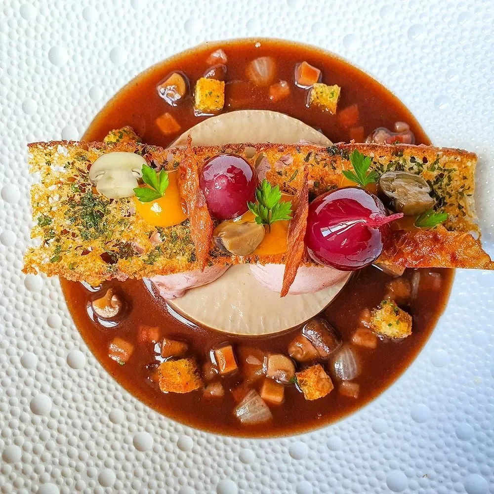 Assiette artistique avec un ragoût de viande, garnie de croûtons, de cerises, de laurier, de champignons, de sauce et de garnitures vertes.