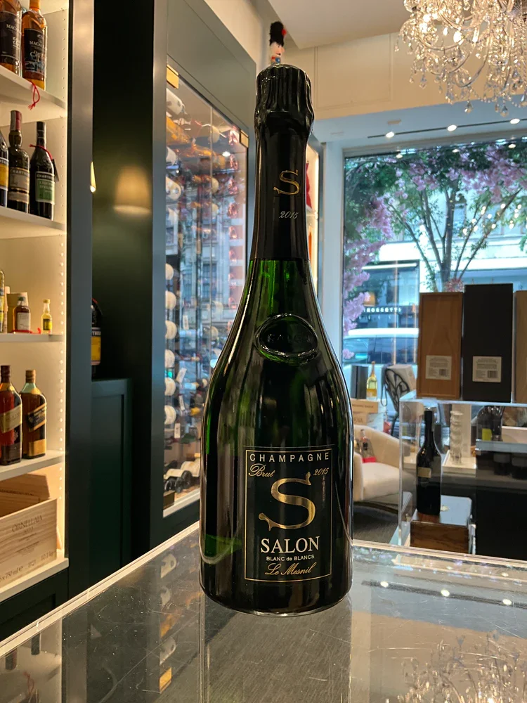 Une grande bouteille de champagne noire avec une étiquette indiquant "Champagne Brut S Salon 2015", posée sur un comptoir en verre dans un magasin ou un salon.