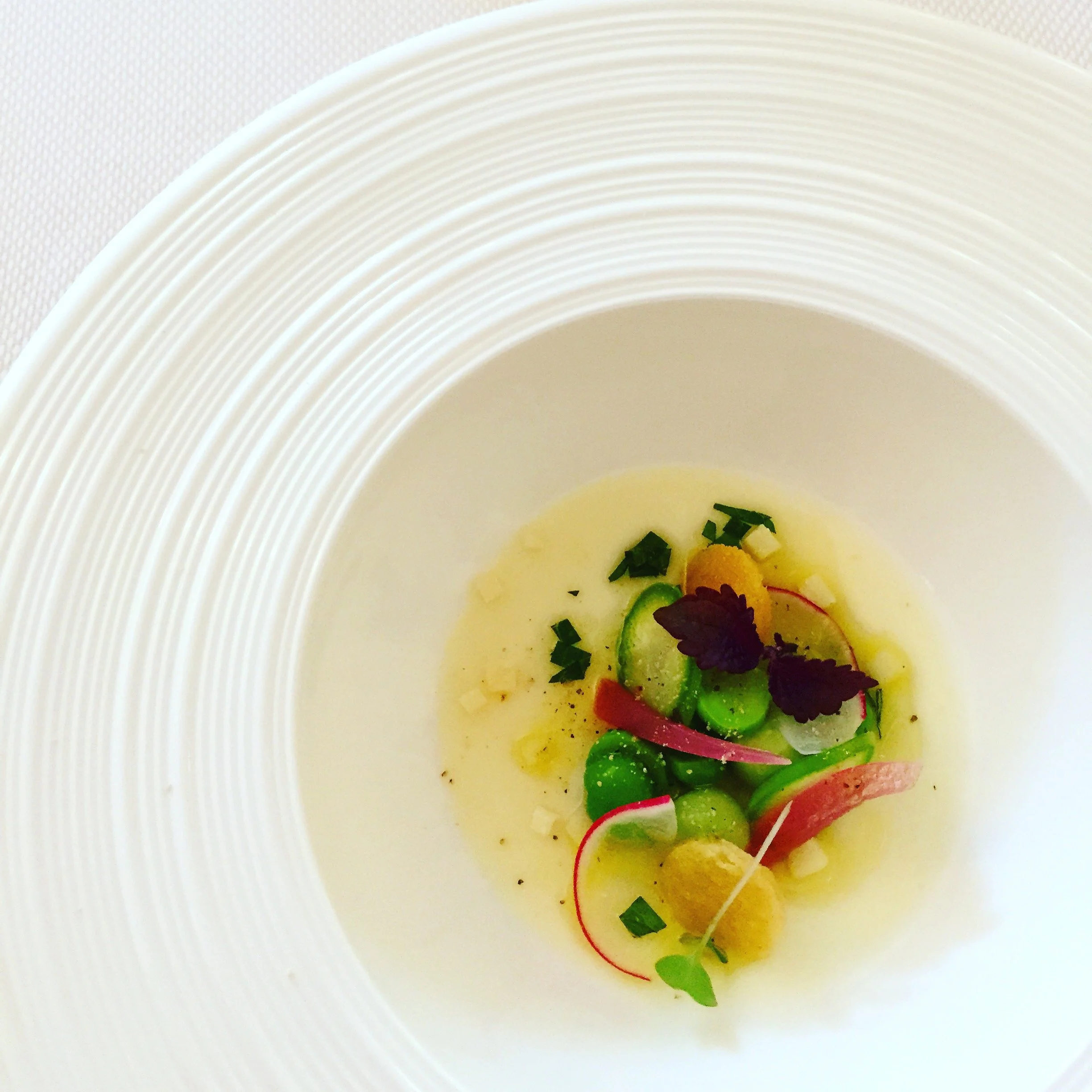 Une présentation de plat gourmet avec des légumes verts, des radis, des herbes et une sauce jaune dans une assiette blanche à bord côtelé.