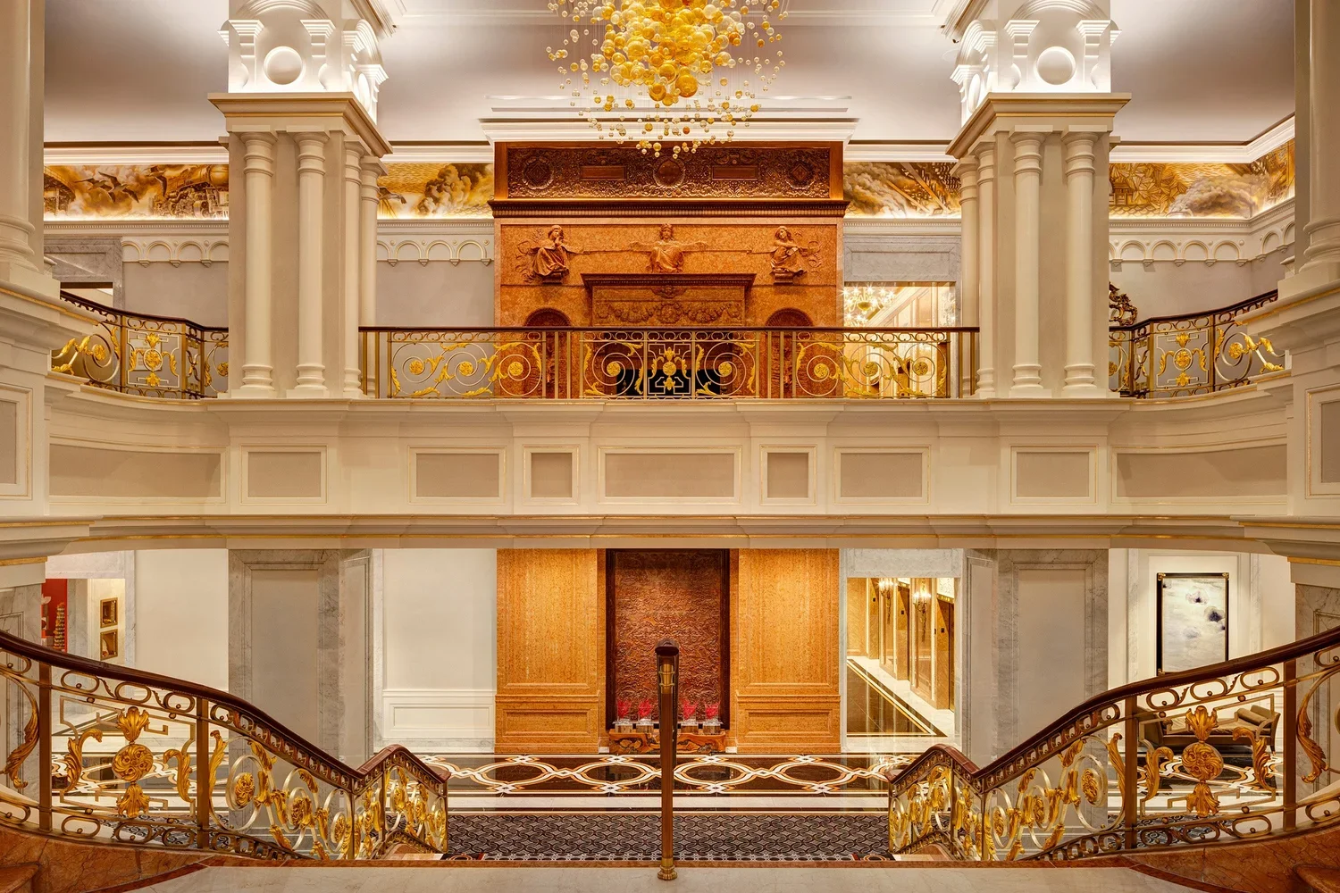 Intérieur luxueux d'un bâtiment avec des balustrades en métal doré, colonnes blanches et décorations en or, plafond orné et sculpture en relief, chandelier, et un grand hall d'entrée avec un motif au sol.