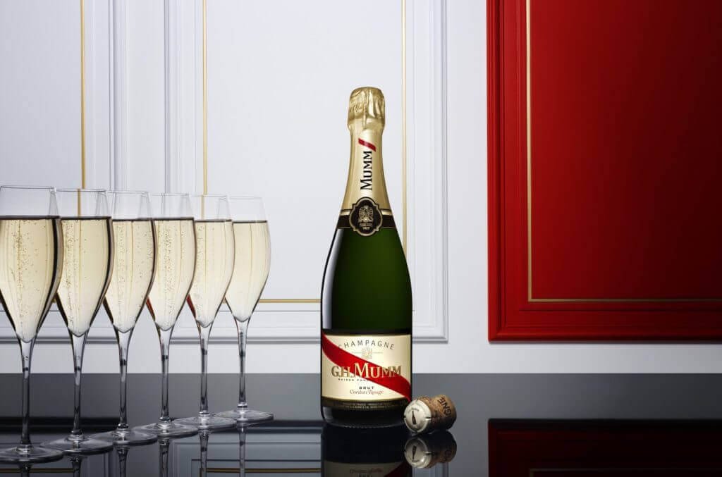 Une bouteille de champagne avec la marque « G.H. Mumm » entourée de six flûtes de champagne pleines dans un décor élégant avec des murs blanc et rouge avec des moulures dorées.