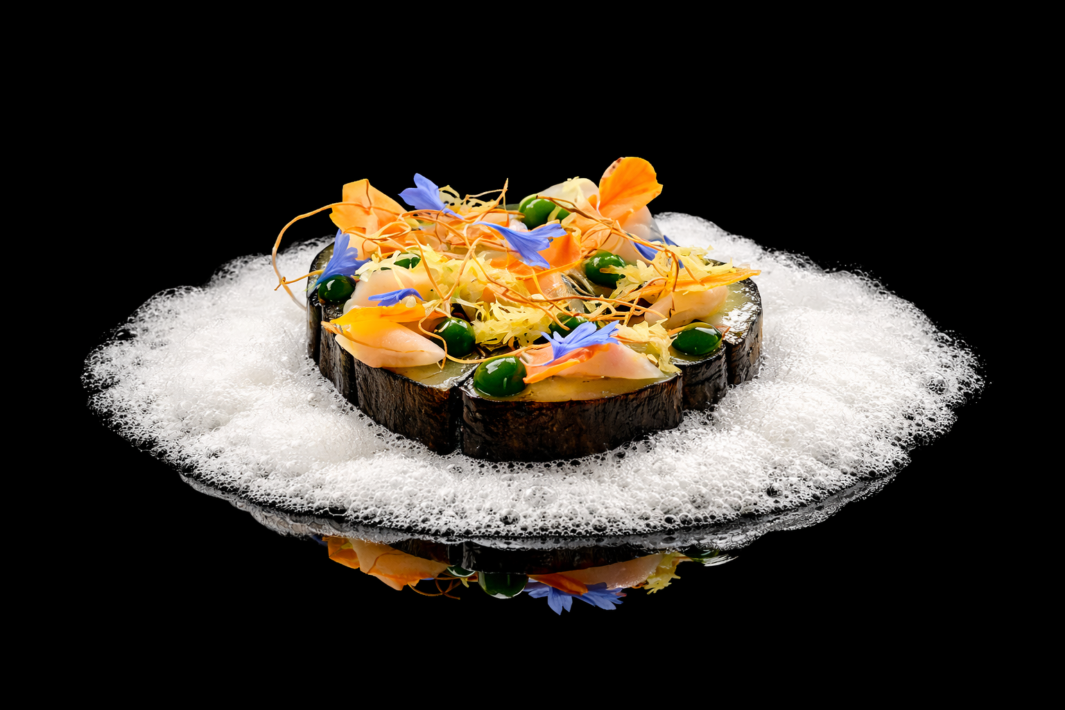 Une présentation gastronomique sur une assiette noire avec un fond noir, comportant une base de bois, surmontée d'une mousse blanche, avec des fleurs comestibles et des garnitures colorées.