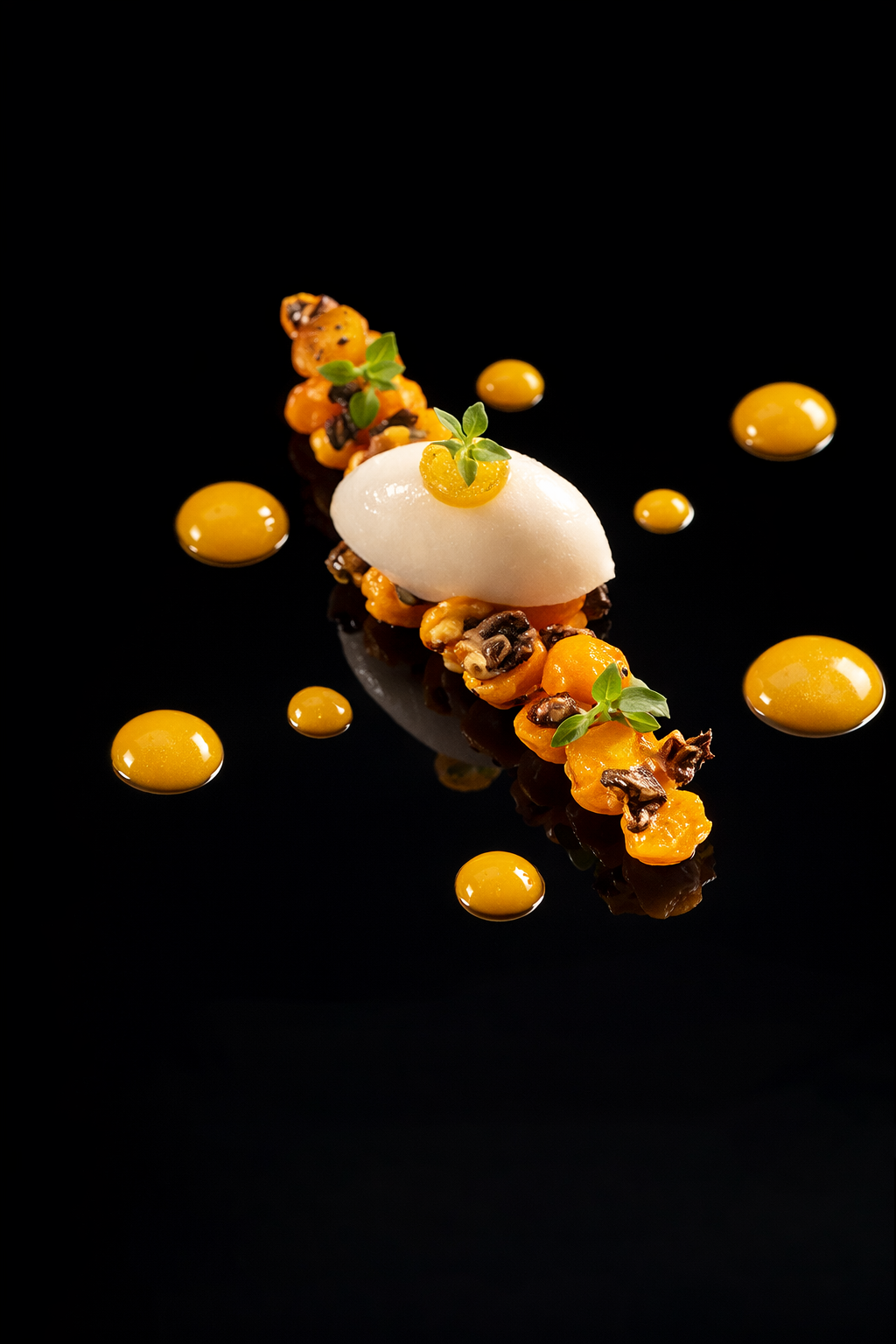Assiette de cuisine gastronomique avec une portion de mousse blanche, des petites tomates jaunes, des micro-pousses, et des gouttes de sauce jaune-orange sur un fond noir.