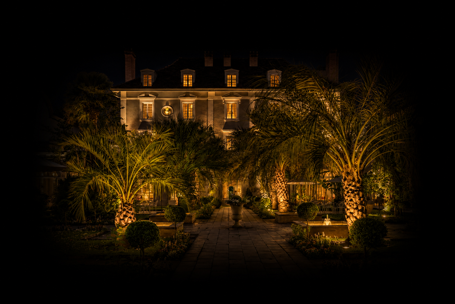 Un bâtiment élégant éclairé la nuit avec un jardin tropical devant, comprenant des palmiers illuminés et un chemin pavé, dans une ambiance chaleureuse.