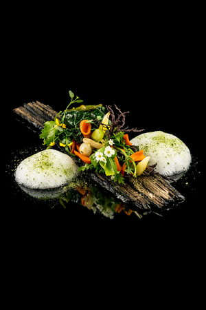 Assiette gastronomique avec deux quenelles, légumes décoratifs et une feuille d'algues, sur une planche en bois noir.