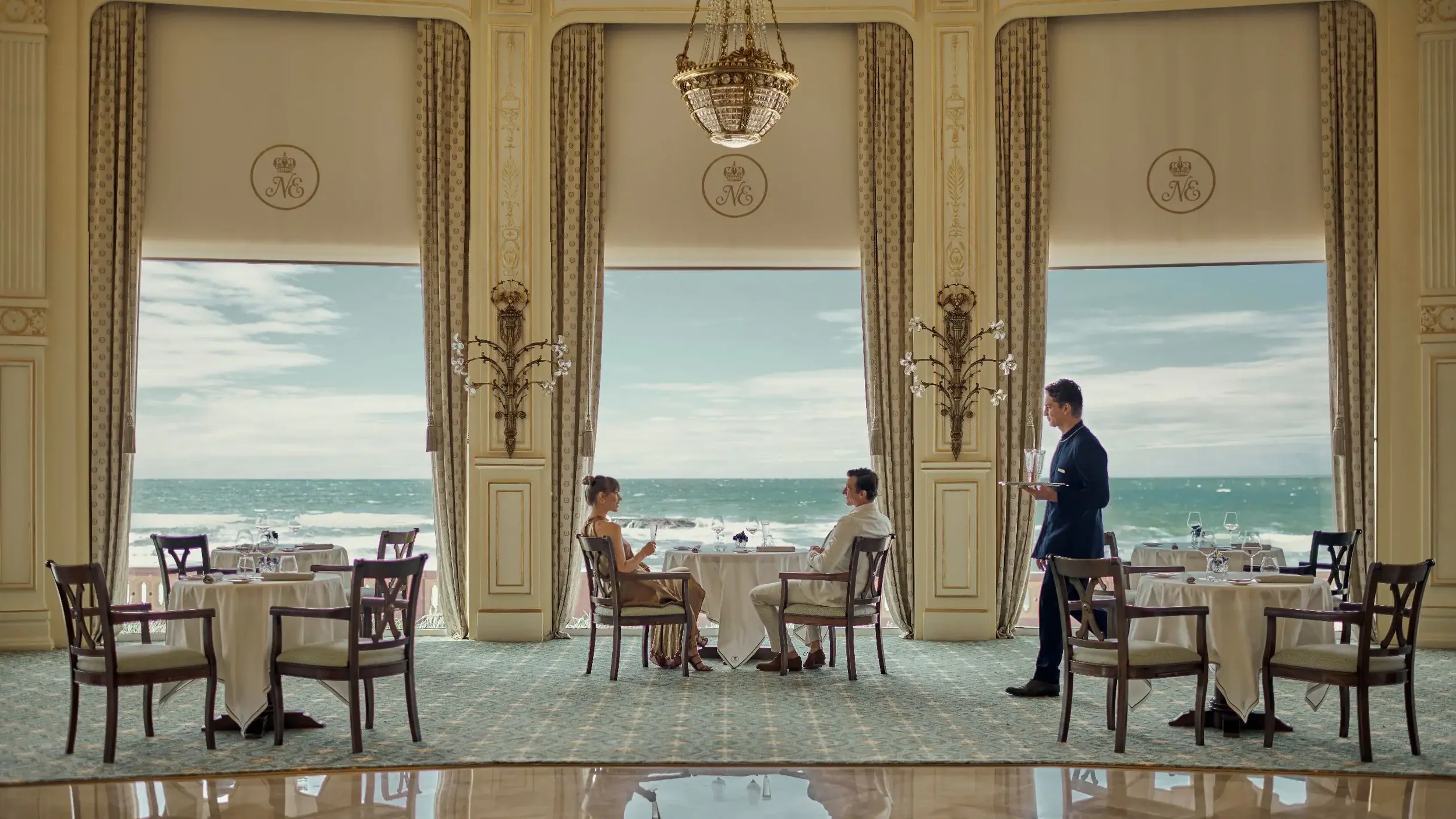 Salle à manger élégante avec vue sur la mer, deux personnes assises à une table, un serveur apportant des plats, grands rideaux, chandeliers décoratifs.