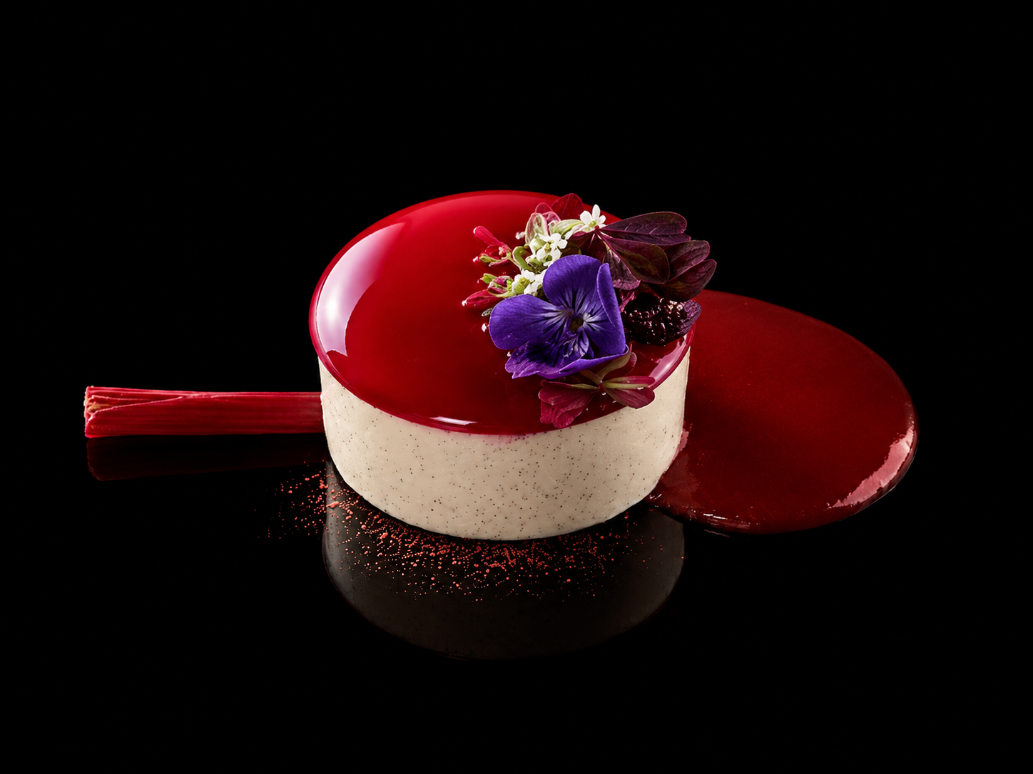 Gâteau rond avec un glaçage rouge brillant, décoré de fleurs violettes, blanches et rouges, posé sur un fond noir.