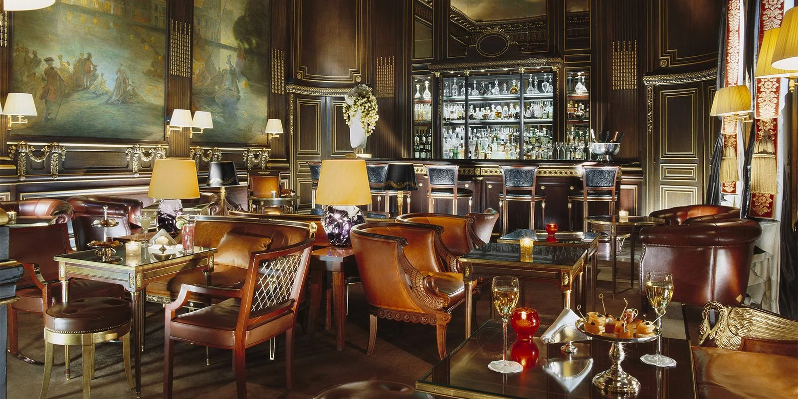 Intérieur d'un bar ou salon élégant avec meubles en cuir, lampes, œuvres d'art, et une étagère remplie de bouteilles et de verres derrière le comptoir.