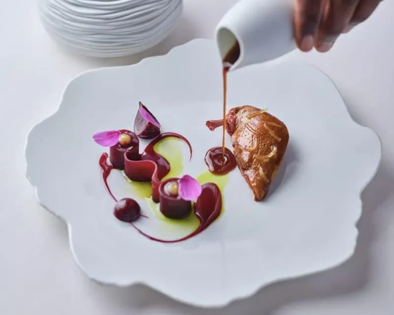 Assiette blanche contenant un plat sophistiqué avec un morceau de viande ou pâtisserie, garniture de purée ou sauce, et décorations avec des fleurs comestibles et des sauces colorées, avec un person en train de verser une sauce brune.
