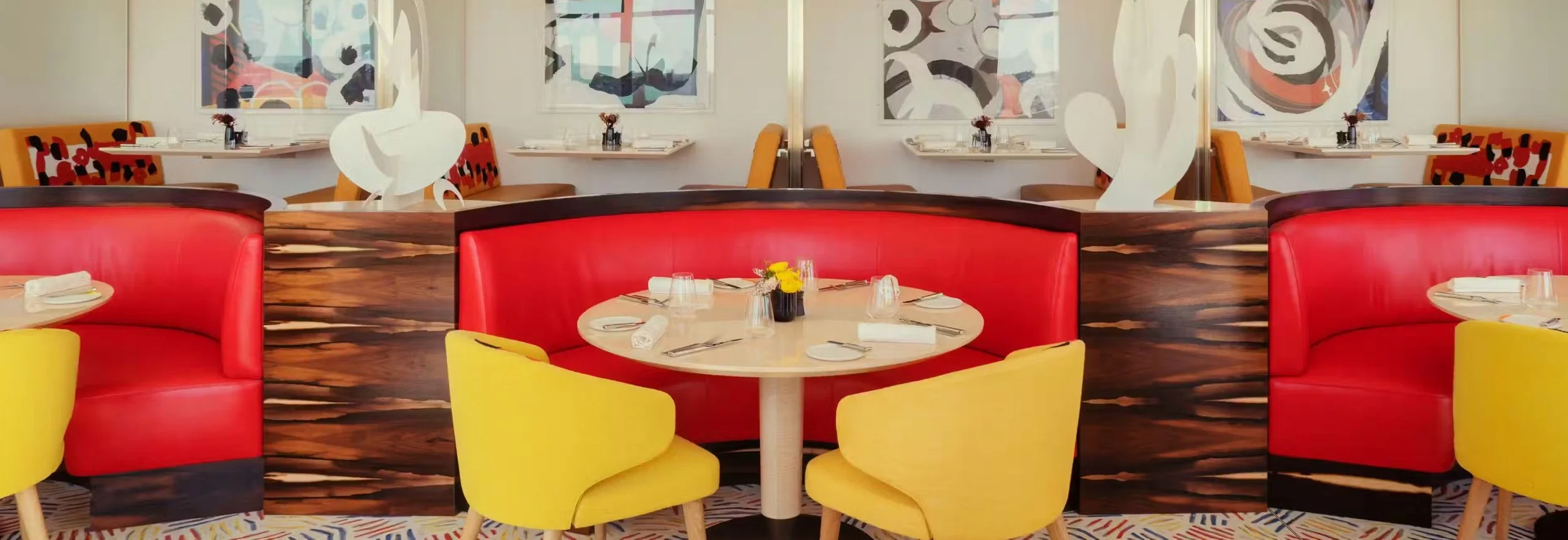 Restaurant avec banquettes rouges et jaunes, tables dressées avec assiettes, couverts, verres et fleurs, décor moderne et artistique sur les murs.